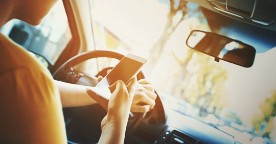 woman sitting in the driver's seat with one hand on the steering wheel and the other hand holding a cellphone