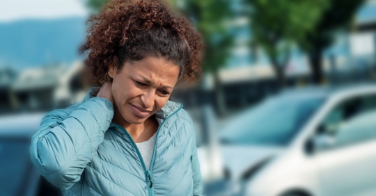 woman holding her neck with a pained facial expression standing in front of a small car with a smashed bumper